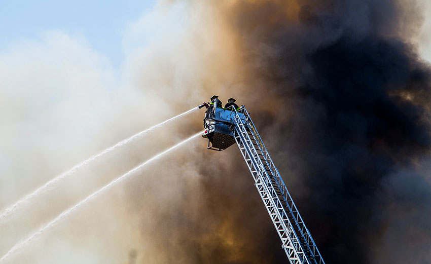 Zwei Feuerwehrmänner auf einer ausfahrbaren Leiter, umhüllt von dunklem Rauch spritzen Wasser aus Schläuchen