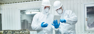 Gefahrstoffbeseitigung: Zwei Männer in weißen Schutzanzügen, blauen Handschuhen, Mundschutz und Schutzbrille stehen vor einer Art Lagerhalle und tippen gemeinsam auf ein iPad