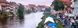 Menüpunkt Kontakt. Ansicht auf Bamberg am Kanal, es sind Menschenmassen, Fahrgeschäfte und Kirchweihbuden auf der rechten Bildhälfte zu sehen, mittig der Fluß und links eine Häuserfront die an Klein Venedig erinnert zu sehen
